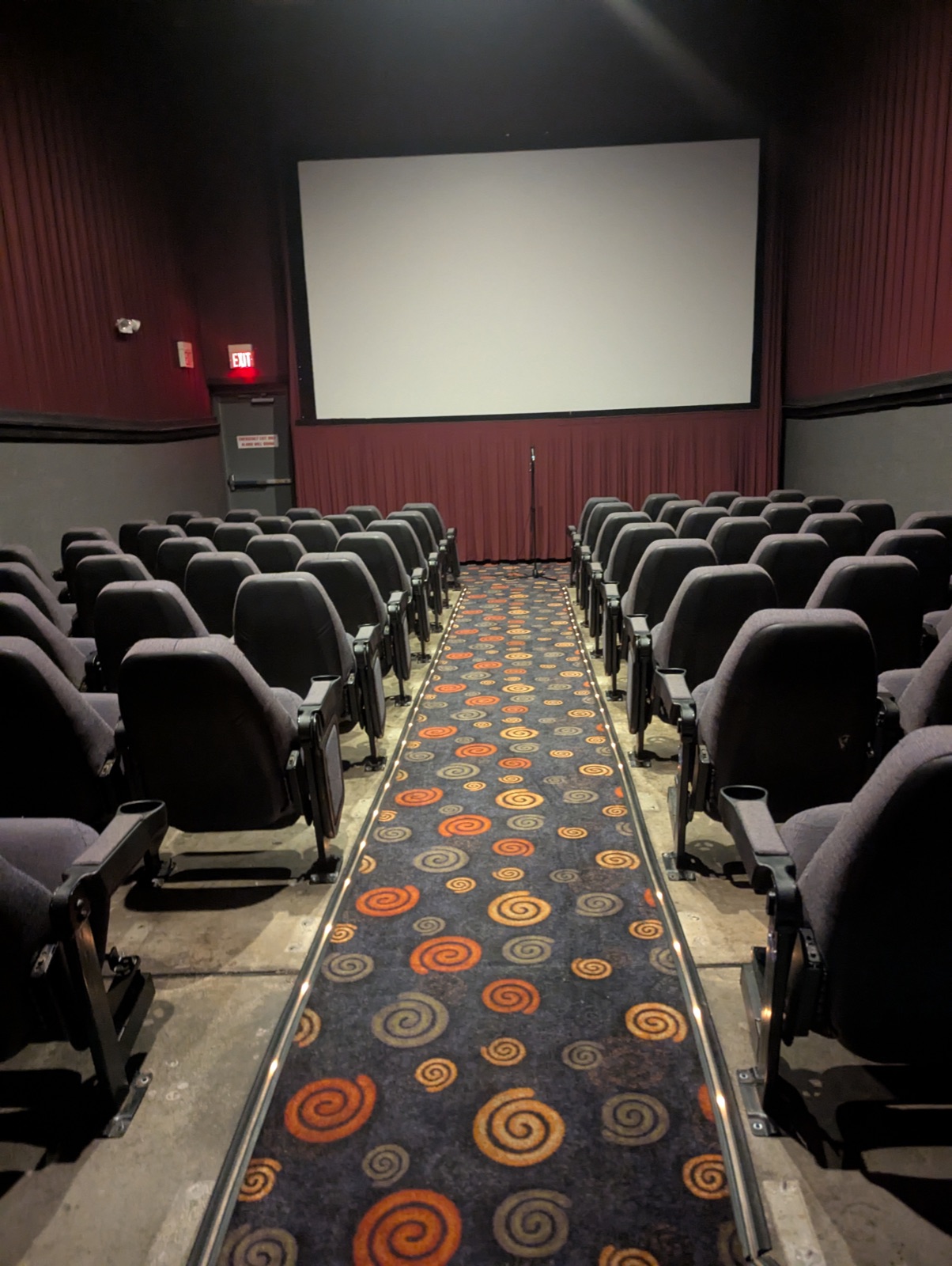 The Esquire's theater empty before doors, rows of grey seats, a swirl-patterned carpet down the aisle and a microphone stand in front of the blank screen