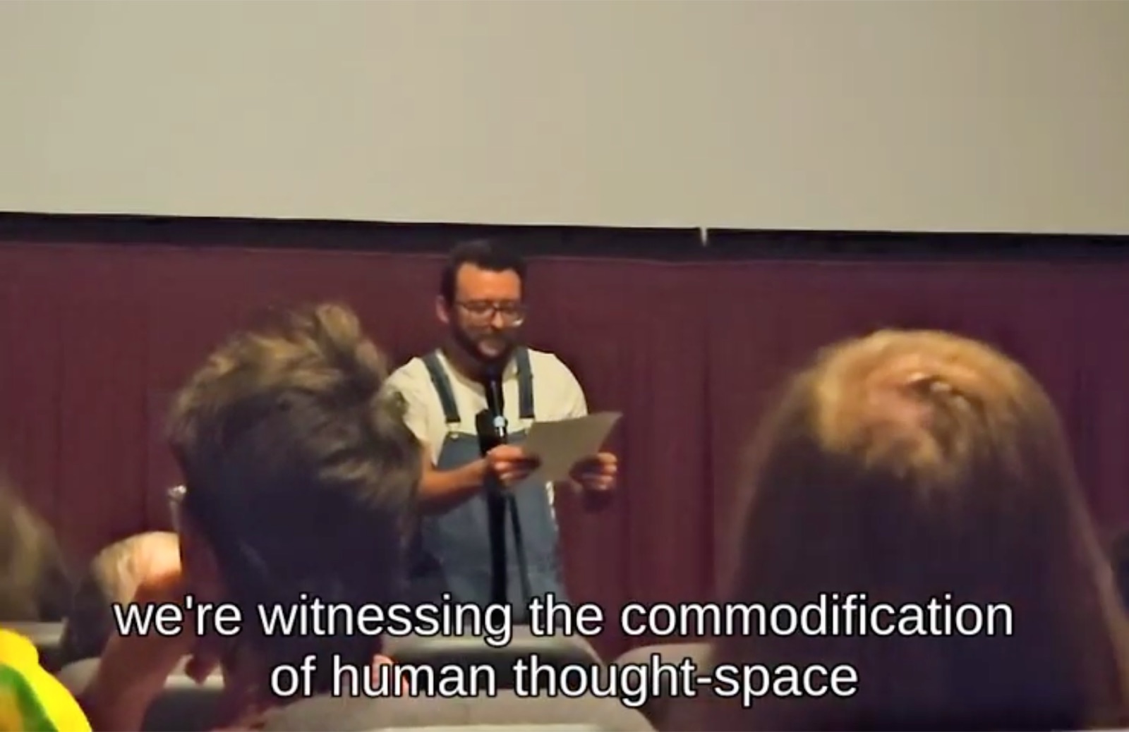 Justin Sovine on stage at the Esquire in overalls, reading the introduction into a microphone in front of the screen, the audience in the foreground